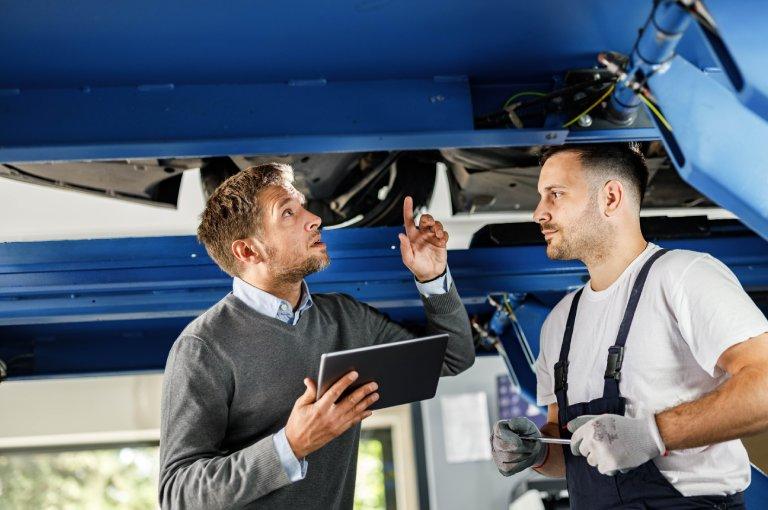 Osoba w swetrze wskazuje mechanikowi na podwozie podniesionego samochodu